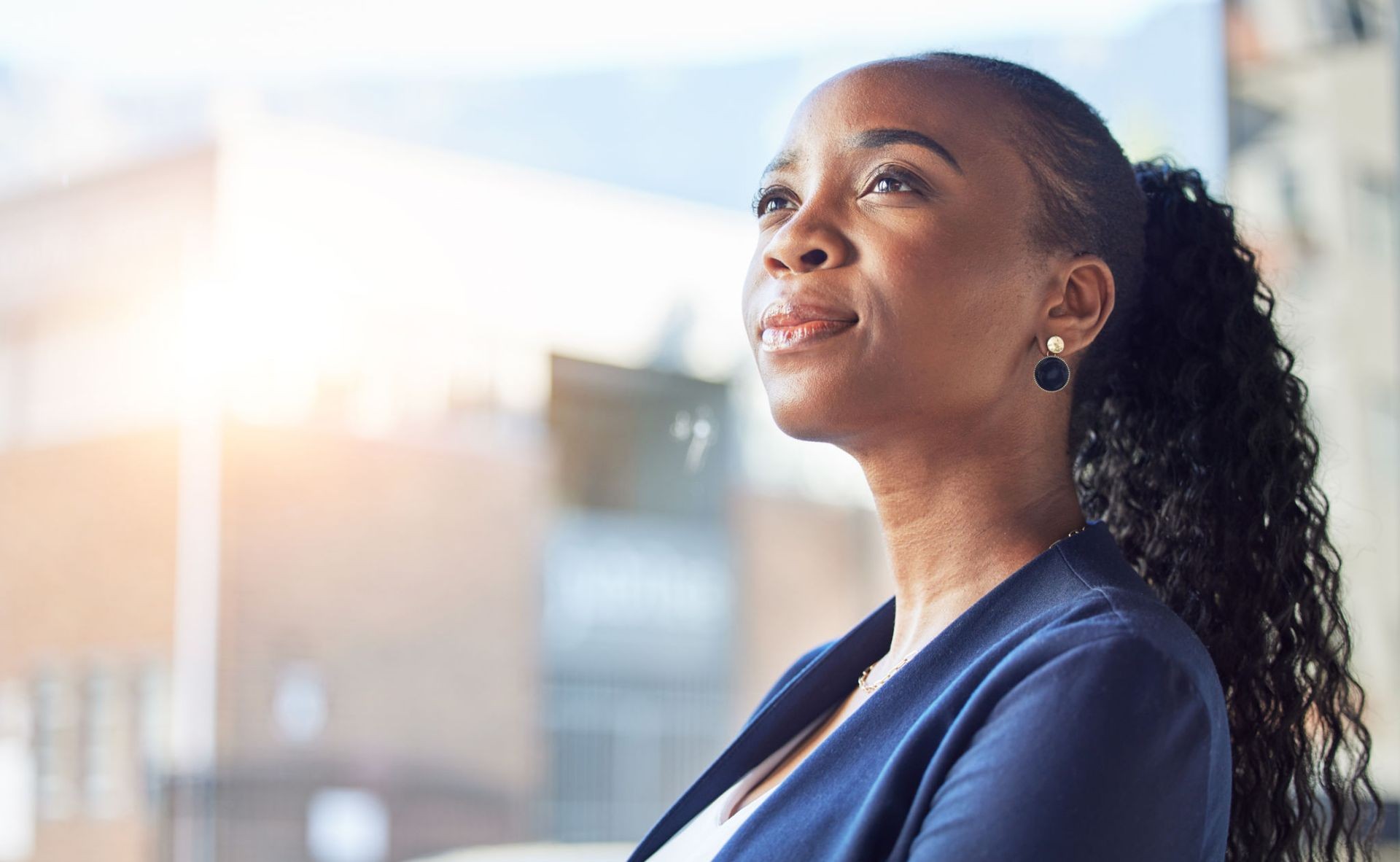 Woman in blue blazer looking upwards with a thoughtful expression, sunlight in the background.