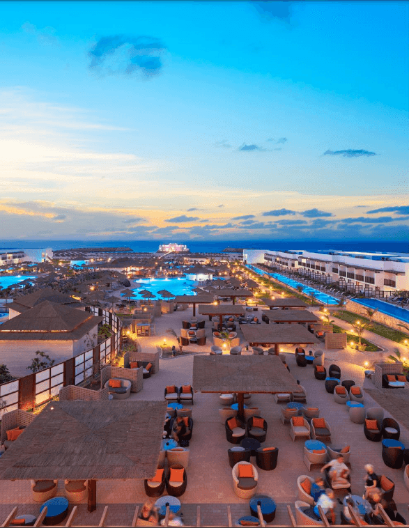 Aerial view of a resort with pools, cabanas, and sunset sky in the background.